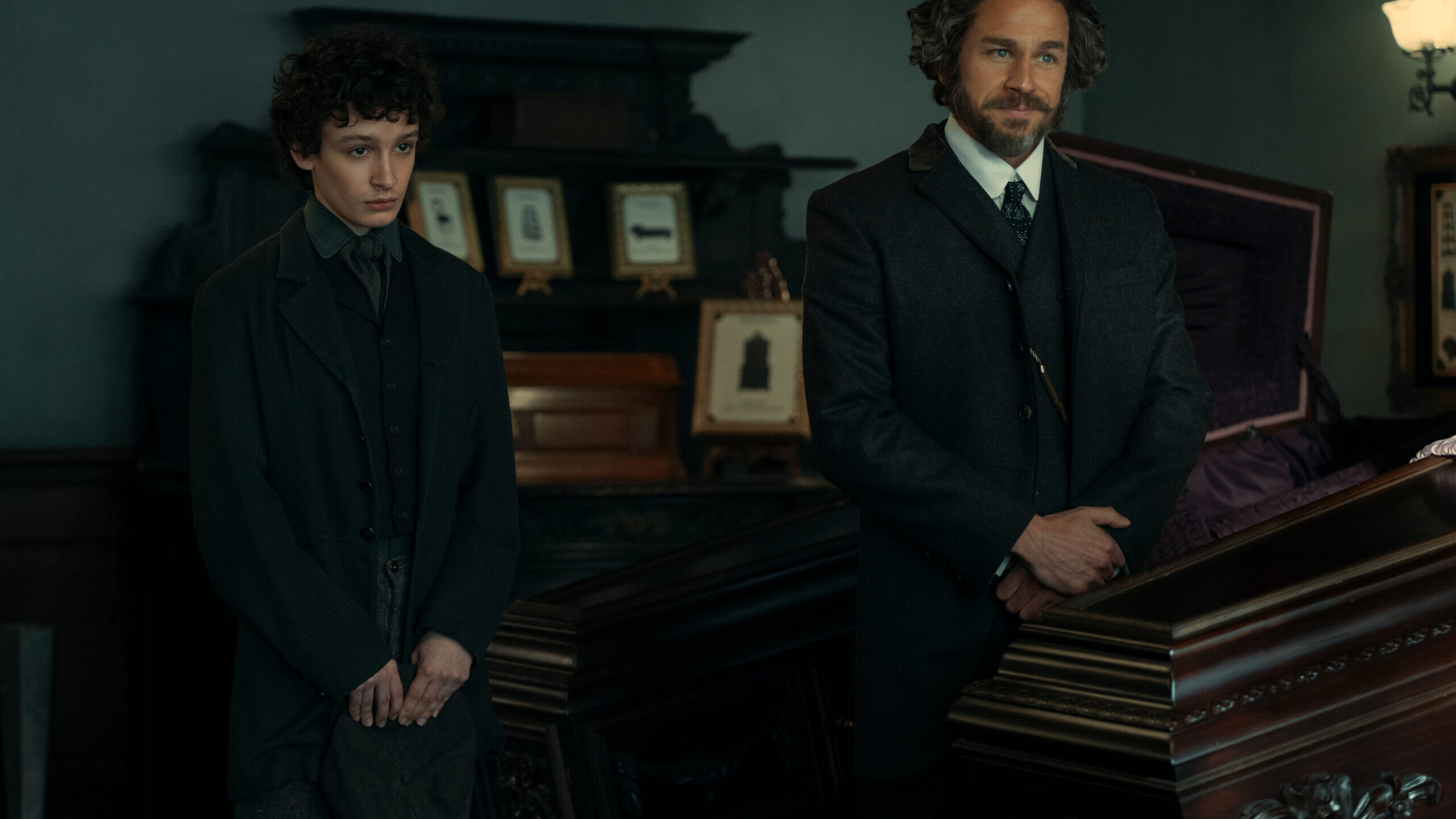 MONSTER_401_Unit_00725RC "Two men in dark, old-fashioned suits stand beside a polished wooden coffin in a dimly lit, ornate room decorated with framed photos, suggesting a 19th-century funeral or wake setting."
