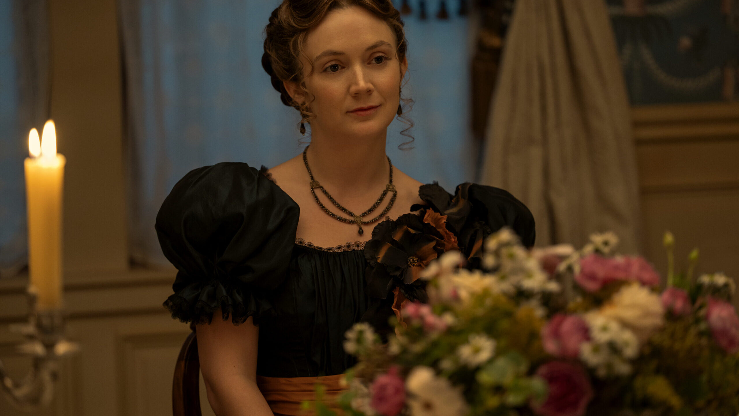 MONSTER_402_Unit_00313RC "Woman in period dress sits at a candlelit table with a bouquet of flowers in an elegant, warmly lit historical room with ornate curtains and soft background decor."
