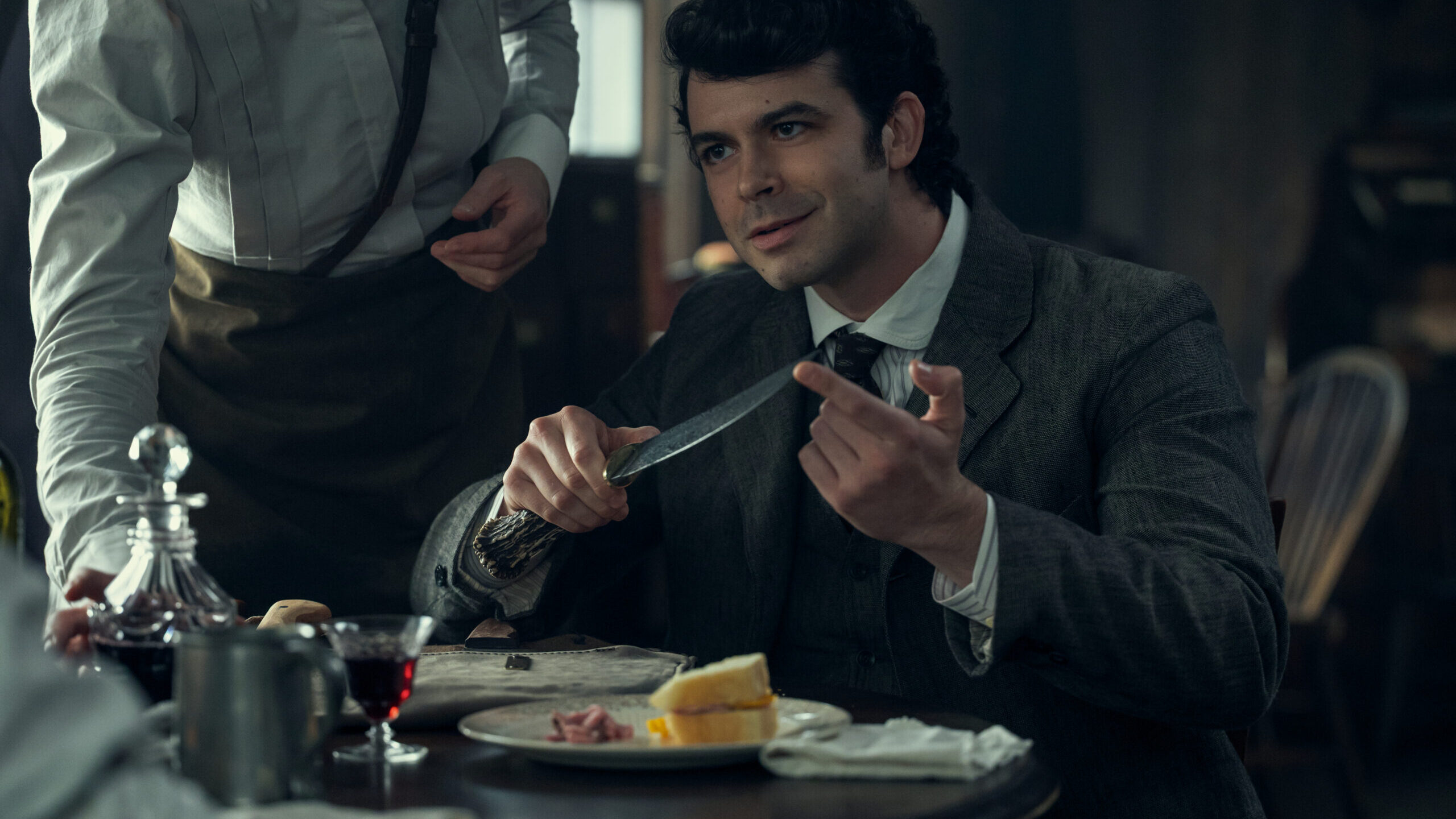 MONSTER_403_Unit_01105RC "Man in vintage suit sits at a table in a dimly lit restaurant holding a knife, with wine, food, and a server nearby, suggesting an early 20th-century dining scene."