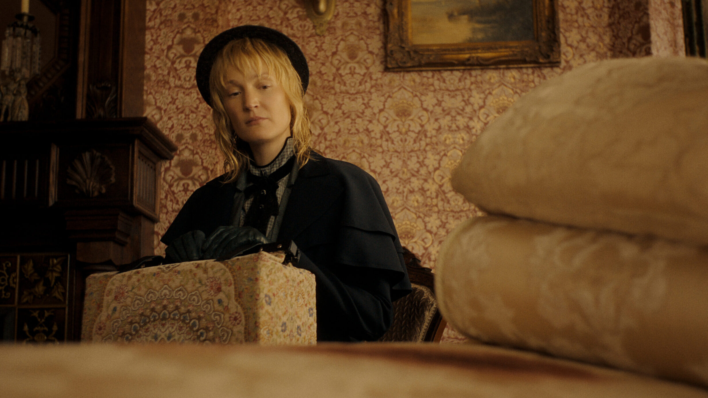 Monster__The_Lizzie_Borden_Story_n_S1_E1_00_19_02_10R "A woman in vintage clothing and gloves sits indoors, holding a patterned box. The room has ornate wallpaper, a painting, and folded linens in the foreground, suggesting a historical or period setting."