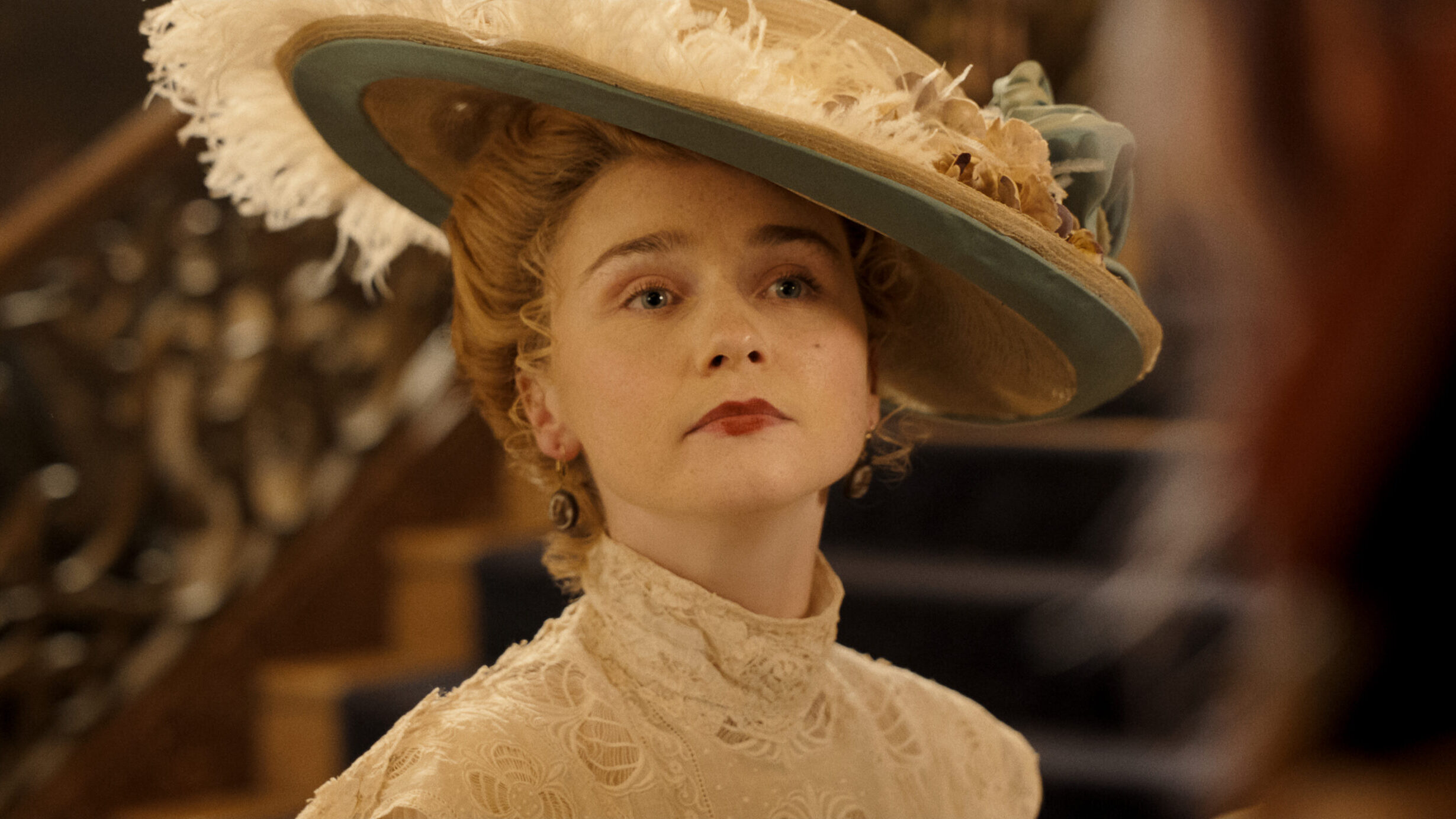 Monster__The_Lizzie_Borden_Story_n_S1_E2_00_17_28_02R "Woman in vintage Edwardian attire and large feathered hat stands indoors near ornate staircase, looking serious and composed, with soft lighting highlighting her intricate lace dress."